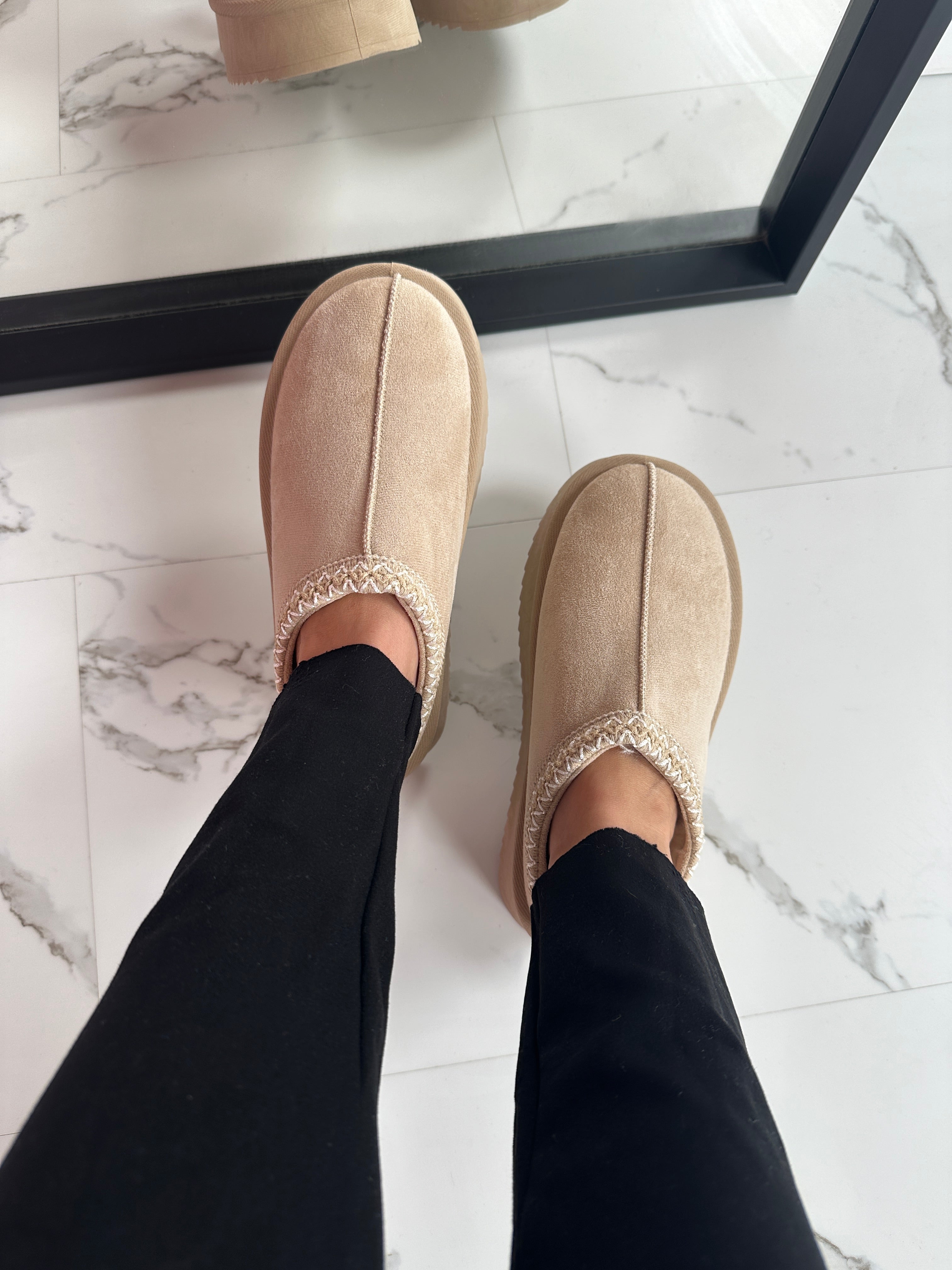 Beige hjemmesko i ruskind med flettet kant, set på marmorflisegulv. Komfortable og stilfulde indendørssko til afslapning derhjemme.