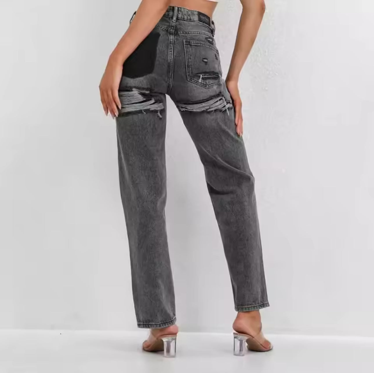 Kvinde iført grå, slidte jeans med huller, set bagfra. Moderne mode, casual stil, trendy denimlook. Høje hæle i gennemsigtig plastik.
