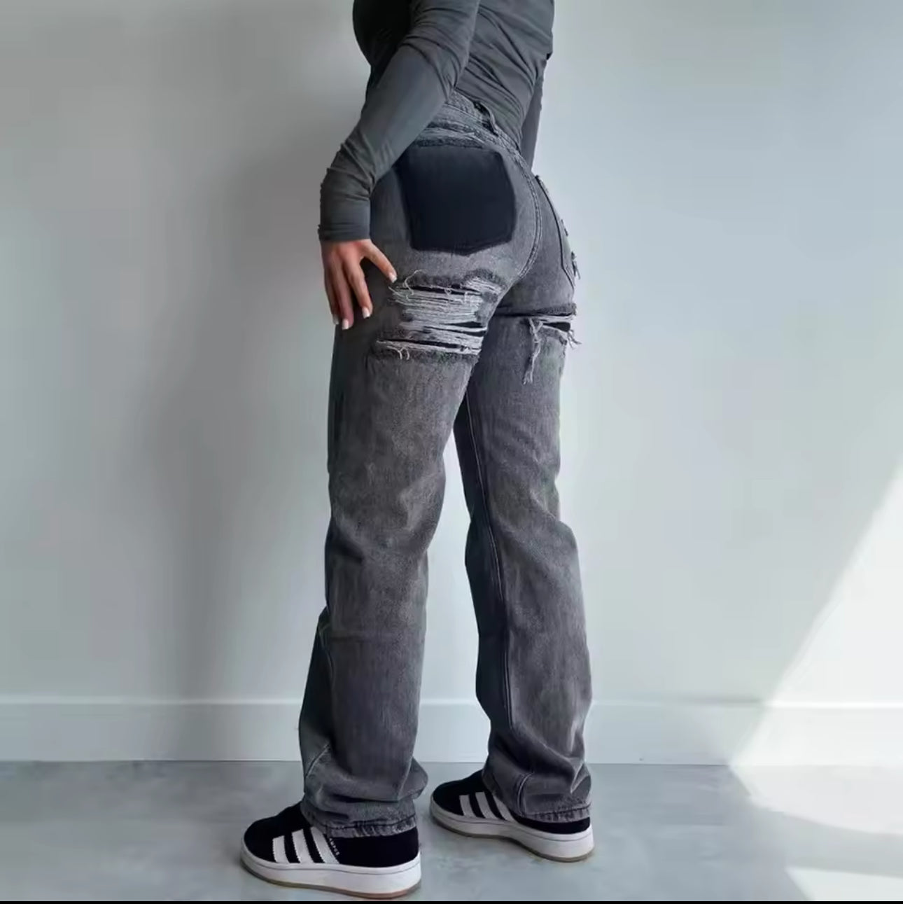 Grå jeans med slidte detaljer og sorte lapper, set bagfra. Person iført grå trøje og sorte sneakers med hvide striber. Moderne streetwear stil.