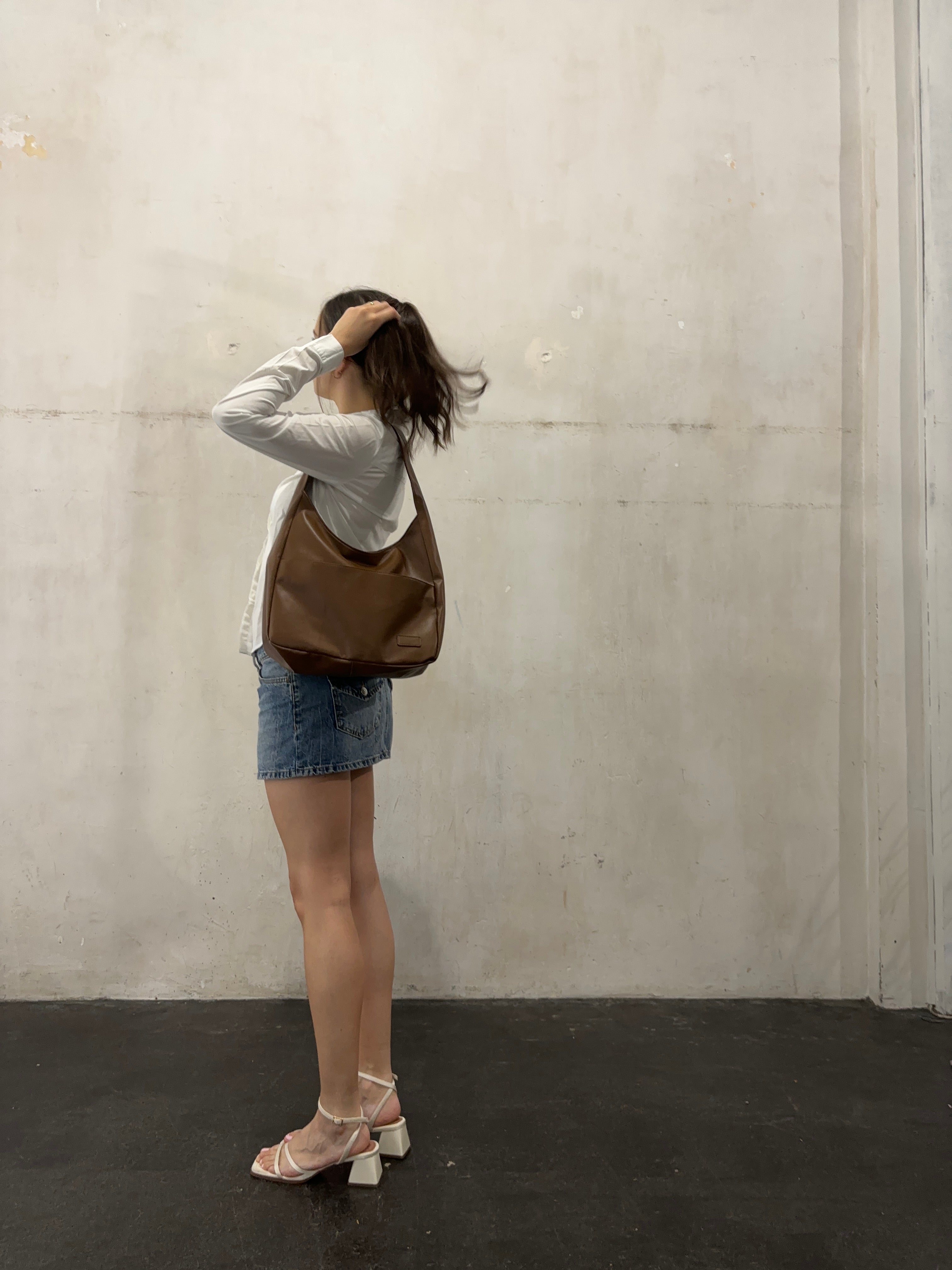 Kvinde i hvid bluse og denimnederdel med brun skuldertaske, poserer mod en rå betonvæg. Mode, casual stil, moderne accessories.