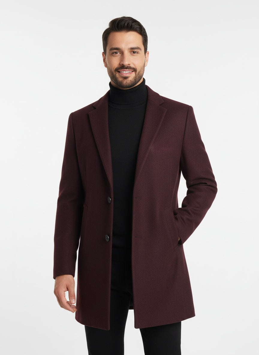 Mand iført bordeaux frakke og sort rullekrave, smilende, stående mod hvid baggrund. Elegant herremode, vinterjakke, stilfuldt look.