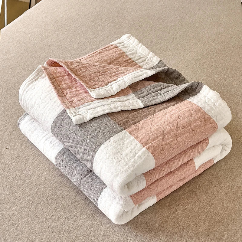 Foldet quiltet tæppe i bløde pastelfarver, herunder lyserød, hvid og grå, på en beige overflade. Perfekt til moderne boligindretning.