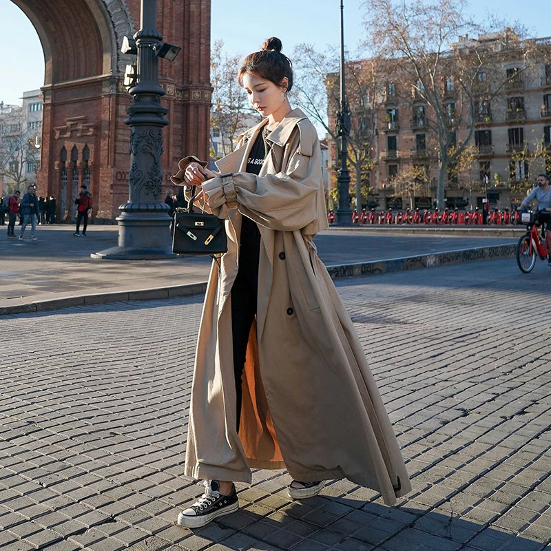 Ung kvinde i lang beige trenchcoat og sorte sneakers, står ved Arc de Triomf i Barcelona. Hun bærer en sort taske og kigger på sin telefon.