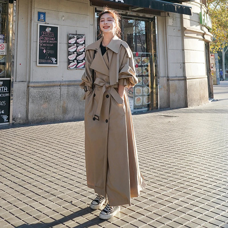 Kvinde i beige trenchcoat og sorte sneakers står smilende på fortovet foran butik. Mode, efterårstøj, street style, byliv.