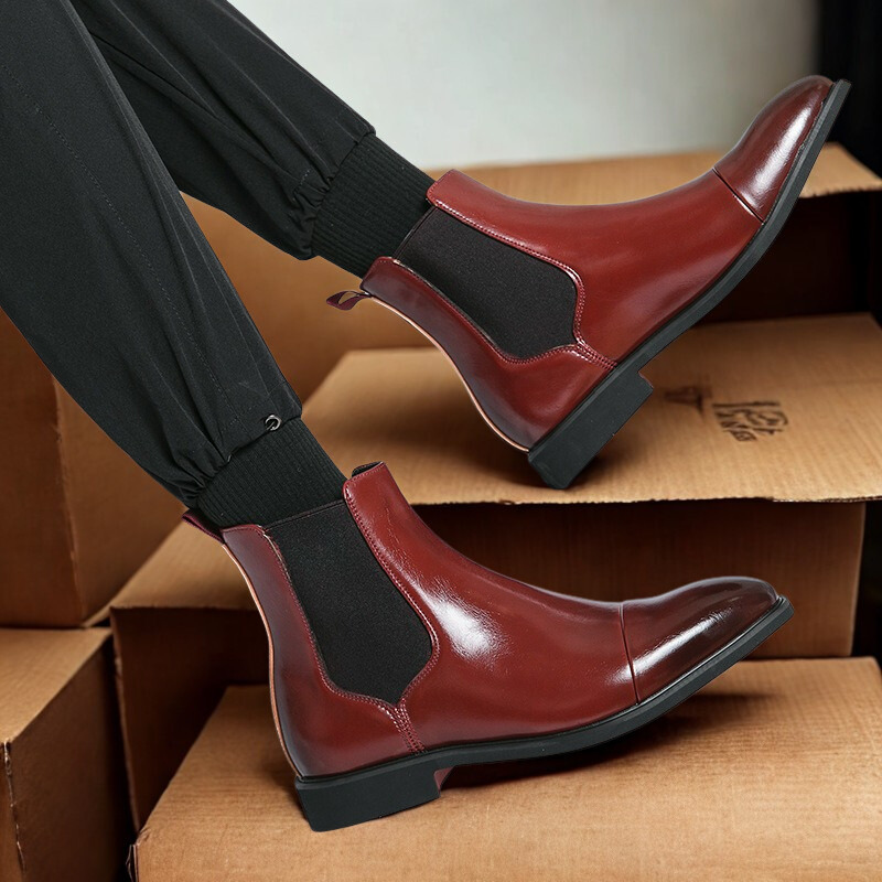 Brune Chelsea-støvler i læder på en person iført sorte bukser, placeret på papkasser. Stilfulde herresko, mode, fodtøj, elegant design.