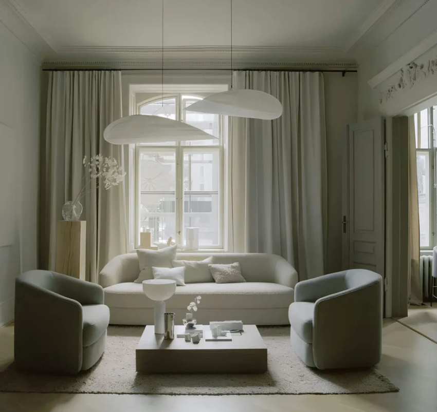 Lyst skandinavisk stue med moderne sofa, to lænestole, minimalistisk sofabord og store vinduer. Elegant belysning og neutrale farver.