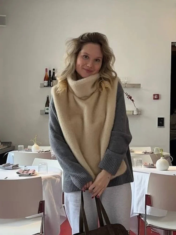 Kvinde i grå sweater og beige tørklæde står i moderne restaurant. Baggrund med hvide borde, stole og vinflasker på hylder.