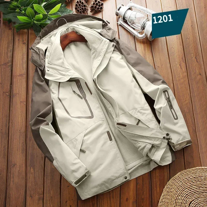 Beige og brun herrejakke med hætte og lynlåse, liggende på trægulv. Udendørsjakke til vandring og friluftsliv. Lanternen ved siden af.