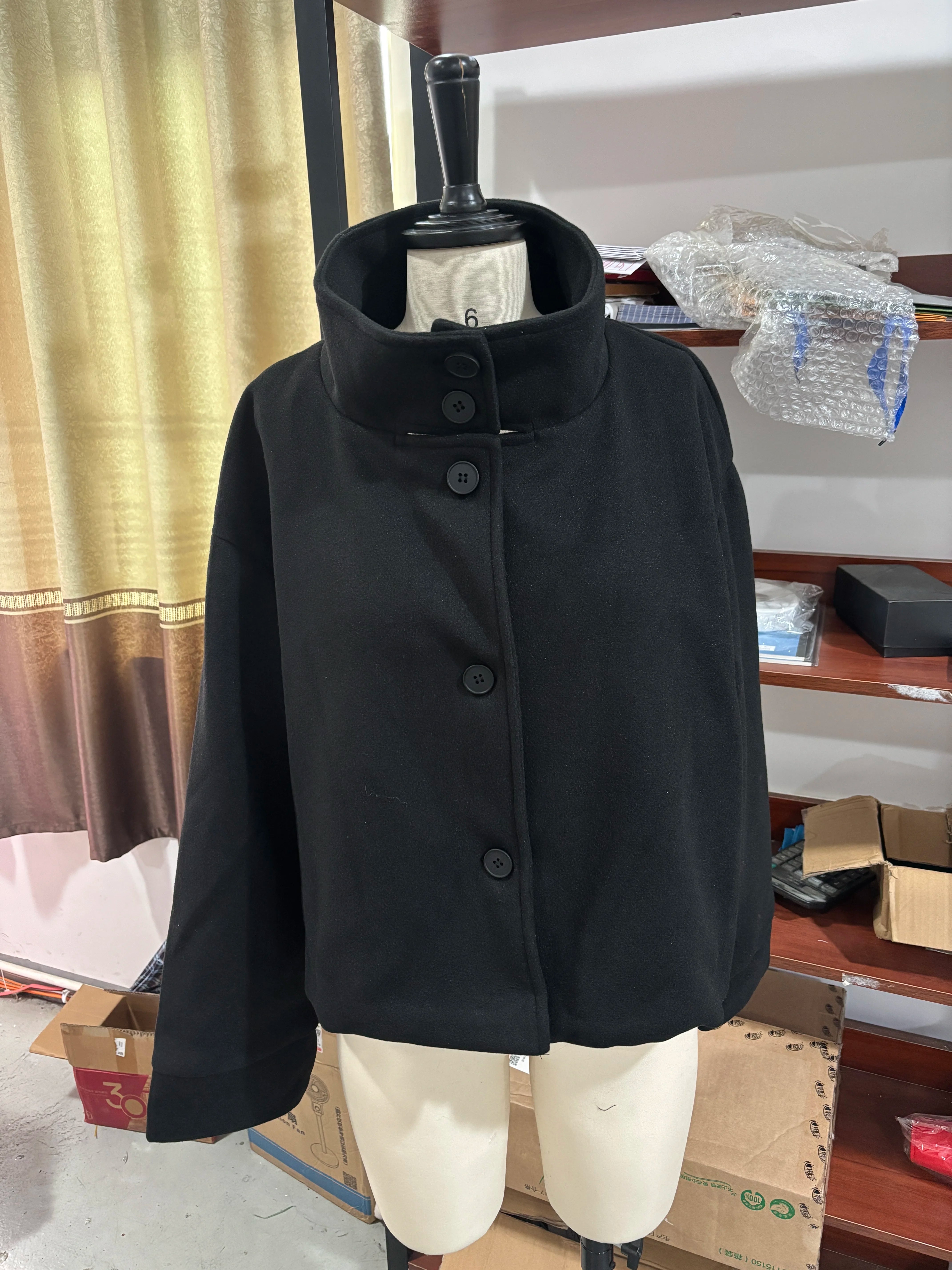 Sort uldfrakke på mannequin, høj krave, knaplukning, moderne design. Perfekt til vintermode. Baggrund med hylder og gardiner.