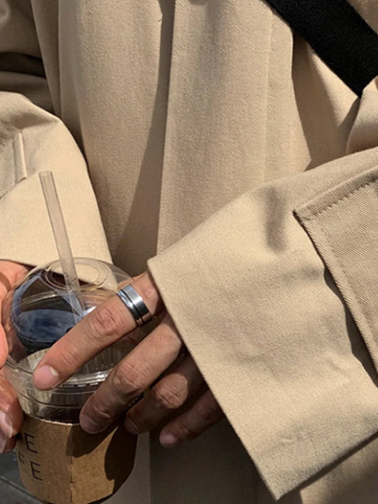 Person iført beige frakke holder en plastikkop med drikkevare og sugerør. Fokus på hænder med ringe. Mode, streetwear, bæredygtig livsstil.