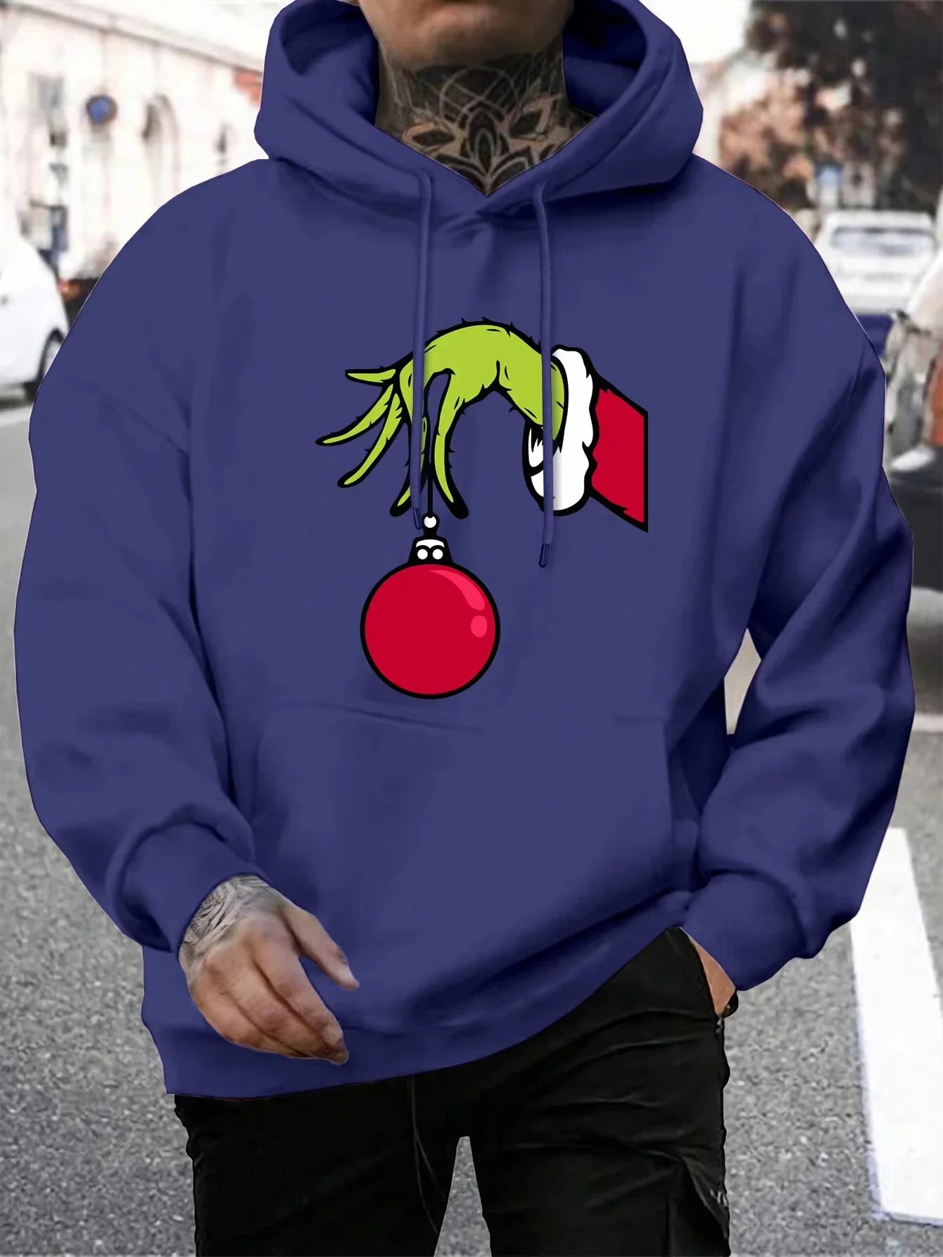 Person iført blå hættetrøje med Grinch-motiv, der holder en rød julekugle. Perfekt til jul, streetwear, unisex mode, vintertøj.