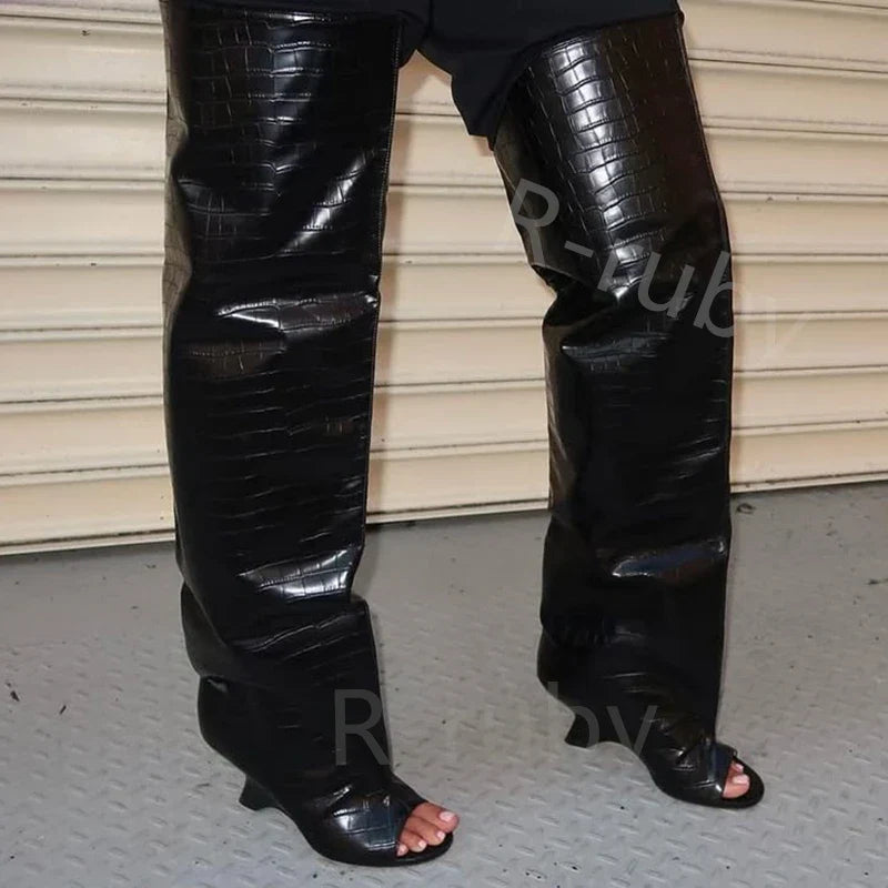 Sort krokodillelæder overknee støvler med åben tå på grå metalgulv foran beige garageport. Trendy fodtøj, luksus mode.