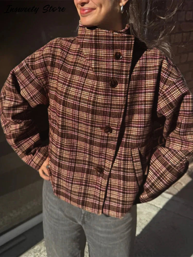 Kvinde iført ternet jakke med store knapper, stående udendørs i sollys. Moderigtig efterårsjakke, brun og rød ternet mønster, casual stil.