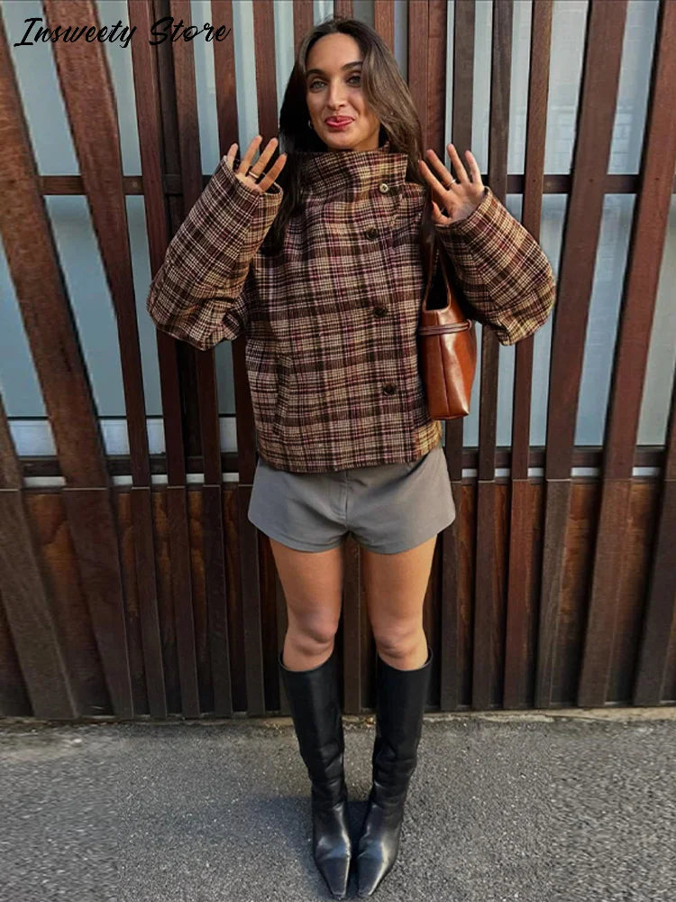 Kvinde i ternet jakke, grå shorts og sorte støvler foran træhegn. Mode, efterårstøj, trendy jakke, stilfuldt look, moderne kvindemode.