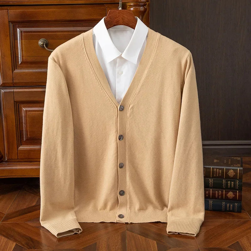 Beige cardigan med knapper over hvid skjorte, hængende på træbøjle foran kommode. Elegant herremode, klassisk stil, efterårstøj.