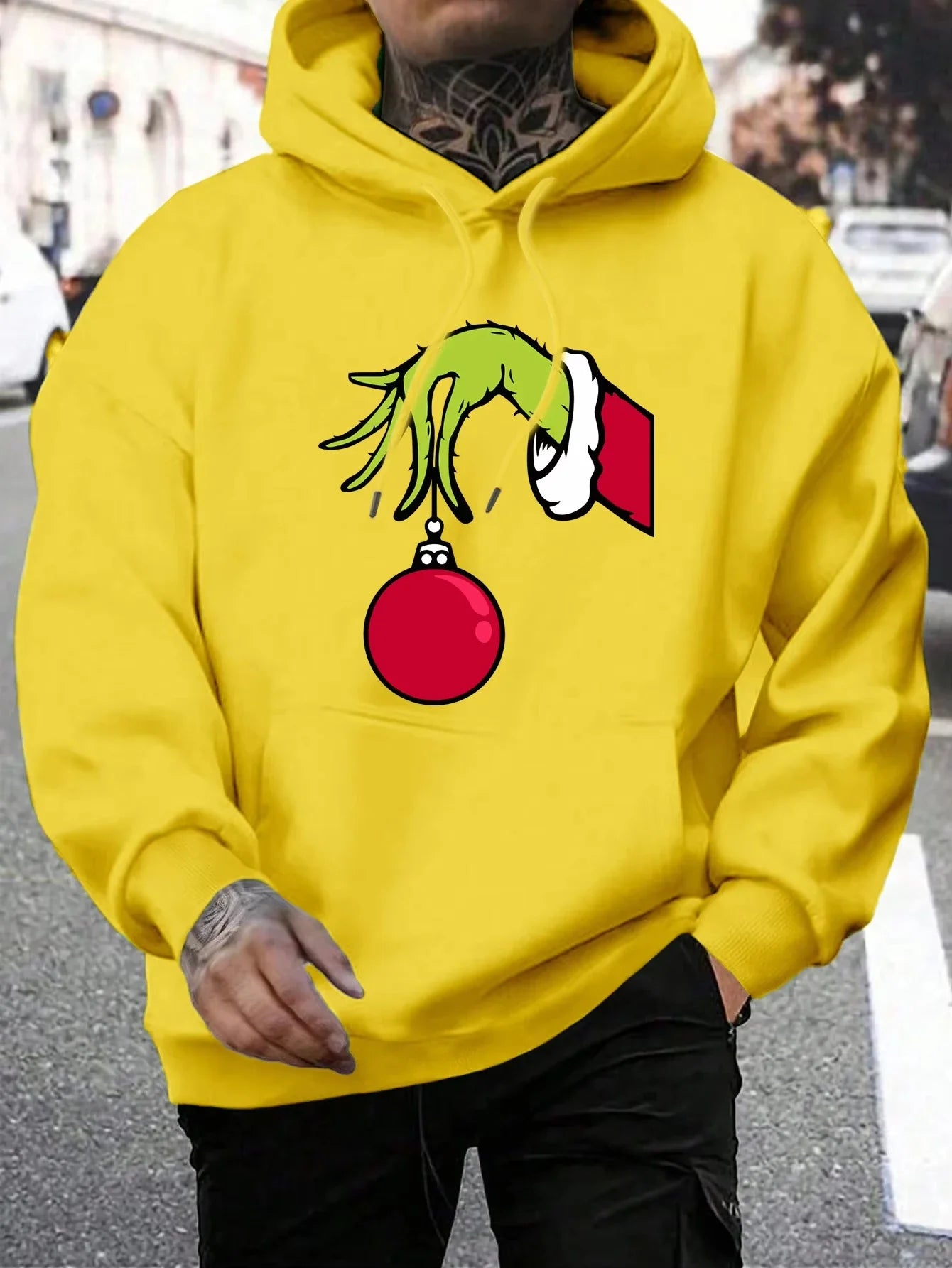 Mand iført gul hættetrøje med Grinch-motiv, der holder en rød julekugle. Streetwear, julemode, unisex hoodie, trendy design.