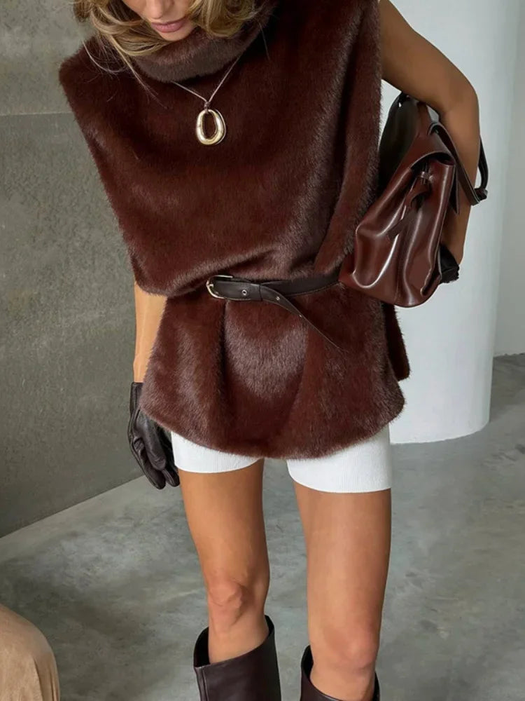 Kvinde iført brun pelsvest, hvid nederdel, sorte handsker og brune støvler. Hun bærer en brun taske og et smykke. Moderne mode, elegant stil.