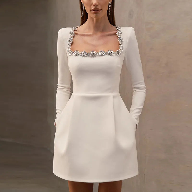 Kvinde i elegant hvid cocktailkjole med firkantet udskæring og blomsterdetaljer, lange ærmer og lommer. Perfekt til festlige lejligheder.