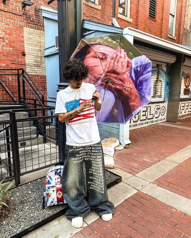 Ung mand med krøllet hår står på gaden med en paraply med et kunstnerisk motiv. Han bærer en hvid T-shirt med amerikansk flag og vide jeans.