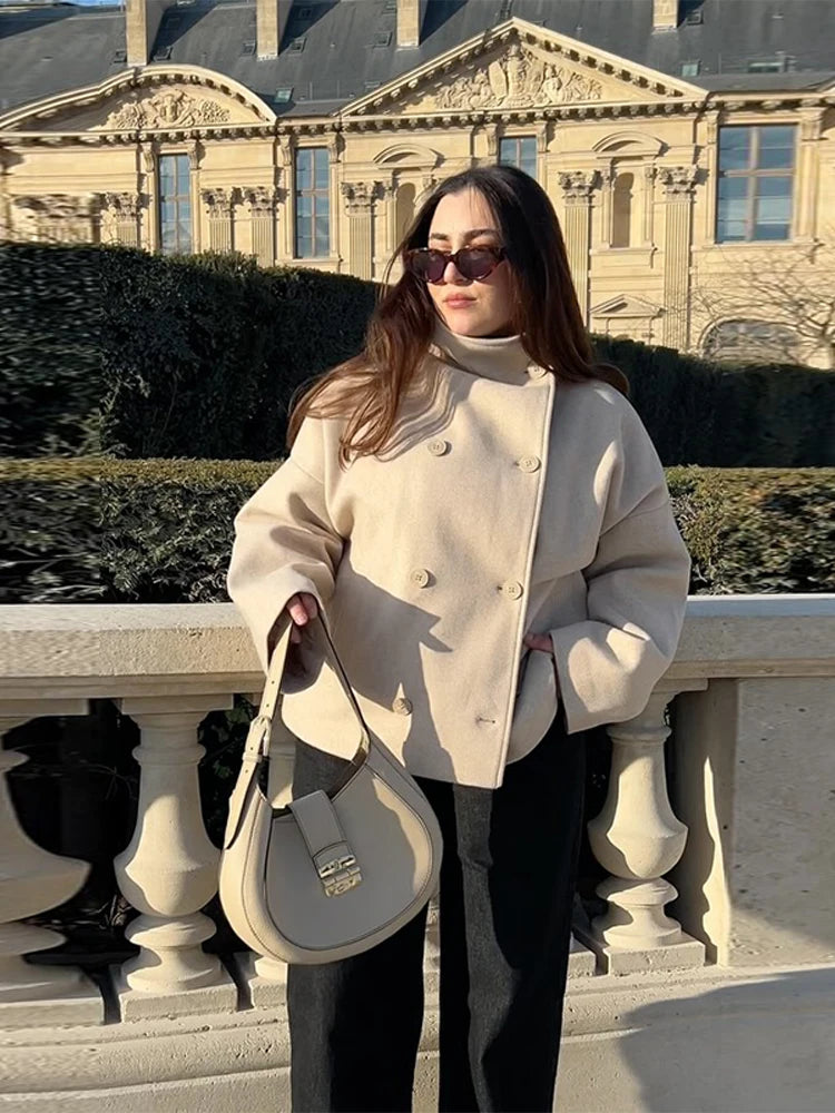 Kvinde i beige frakke og solbriller står foran historisk bygning, holder en stilfuld beige taske. Mode, udendørs, arkitektur, solrig dag.