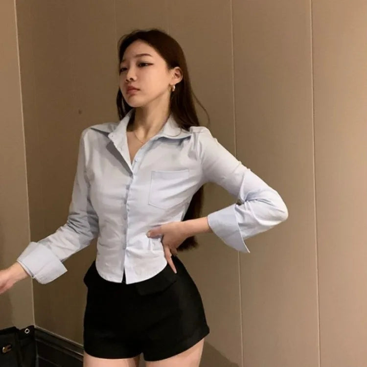 Ung kvinde i hvid skjorte og sorte shorts poserer mod beige væg. Moderne mode, stilfuldt outfit, minimalistisk indretning.