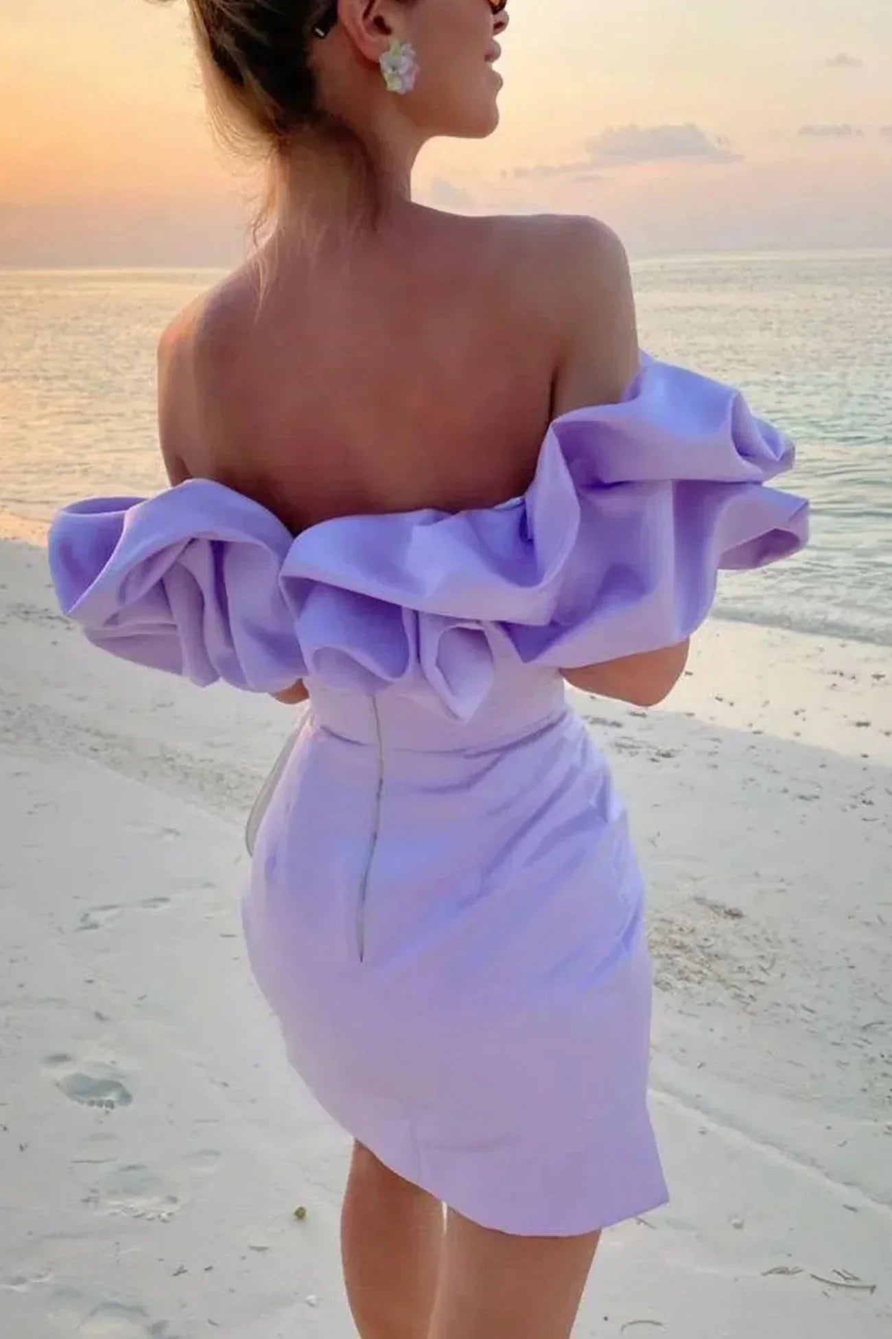 Kvinde i lilla off-shoulder kjole med flæser står på strand ved solnedgang. Elegant sommermode, strandtøj, romantisk aftenstemning.