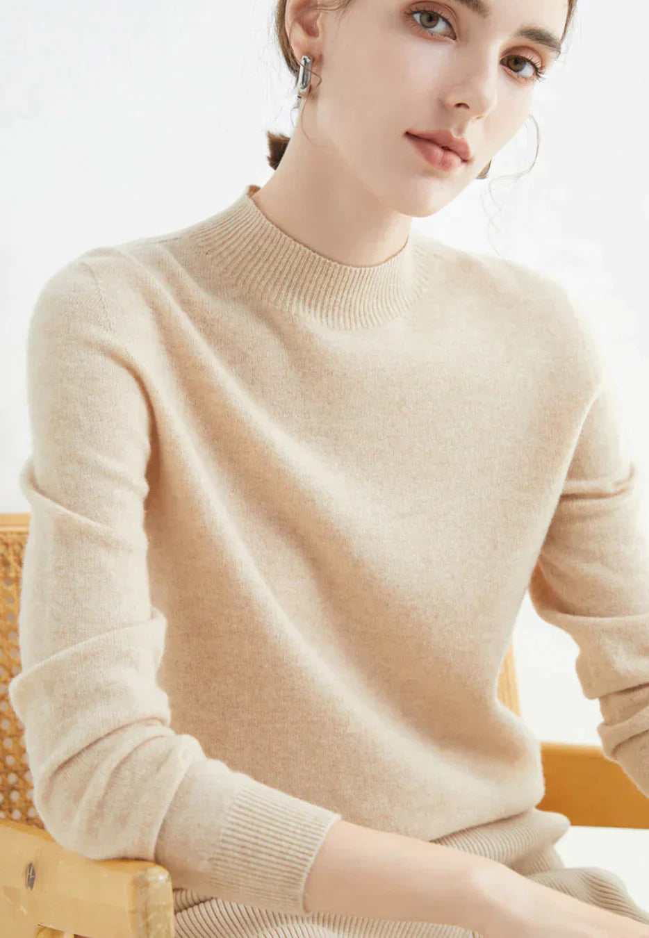 Kvinde iført beige cashmere sweater, sidder på stol. Elegant og minimalistisk mode, perfekt til efterår og vinter. Komfortabelt og stilfuldt design.