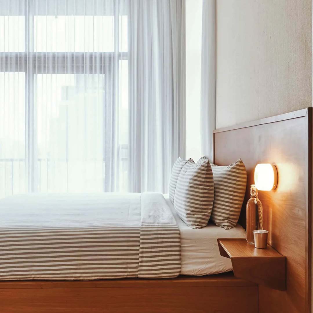 Moderne soveværelse med træramme seng, stribede puder og sengetøj, væglampe og store vinduer med hvide gardiner. Skandinavisk indretning.