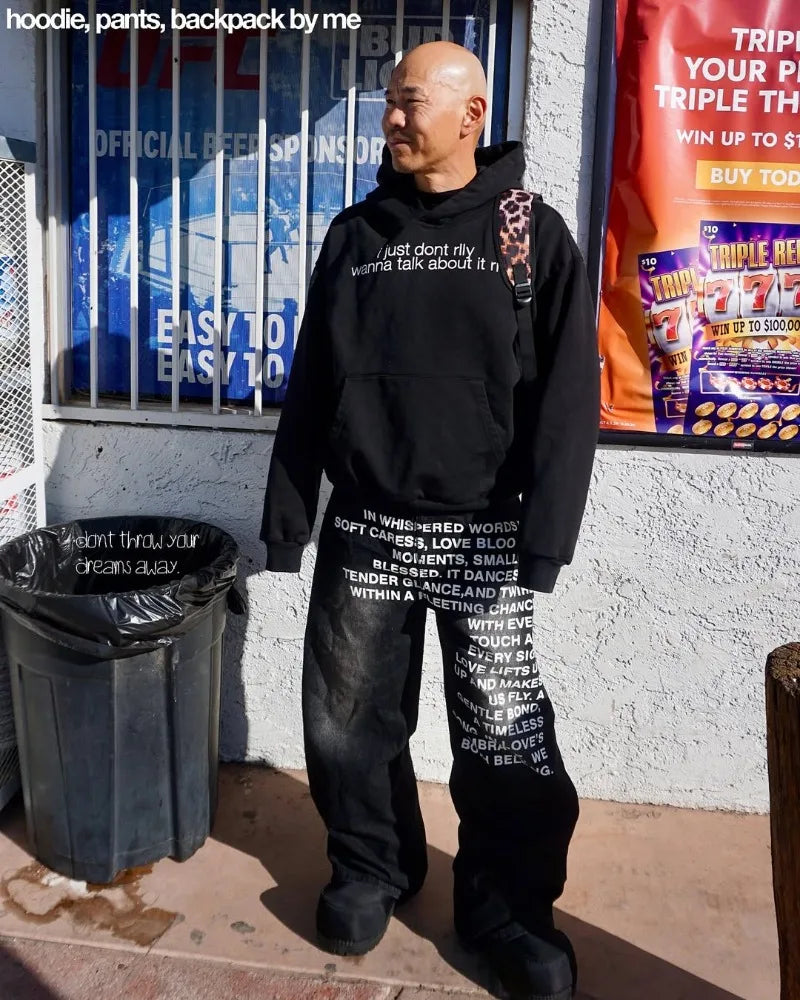 Mand iført sort hættetrøje med tekst, sorte bukser med hvid skrift og rygsæk. Står foran butik med farverige plakater.