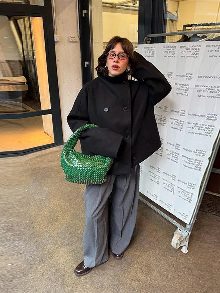 Kvinde i sort jakke og grå bukser med grøn flettet taske. Står foran butiksvindue. Mode, street style, moderne kvindetøj, trendy accessories.