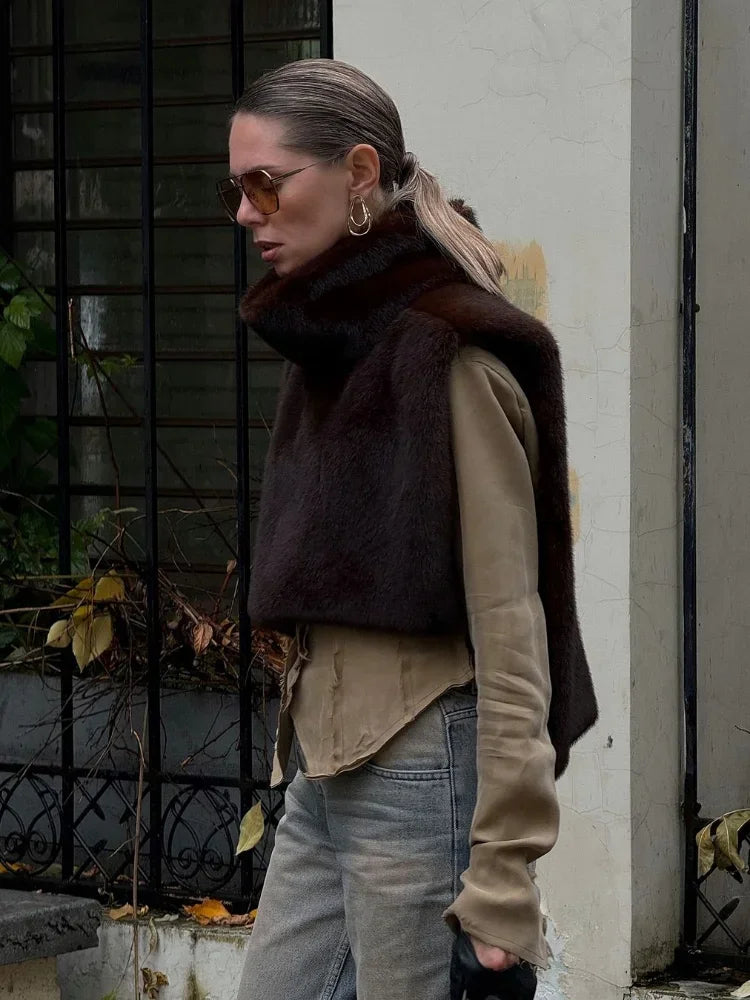 Kvinde iført brun pelsvest, beige skjorte og solbriller, går forbi et gitterhegn. Mode, efterårstøj, stilfuldt look, bymiljø.