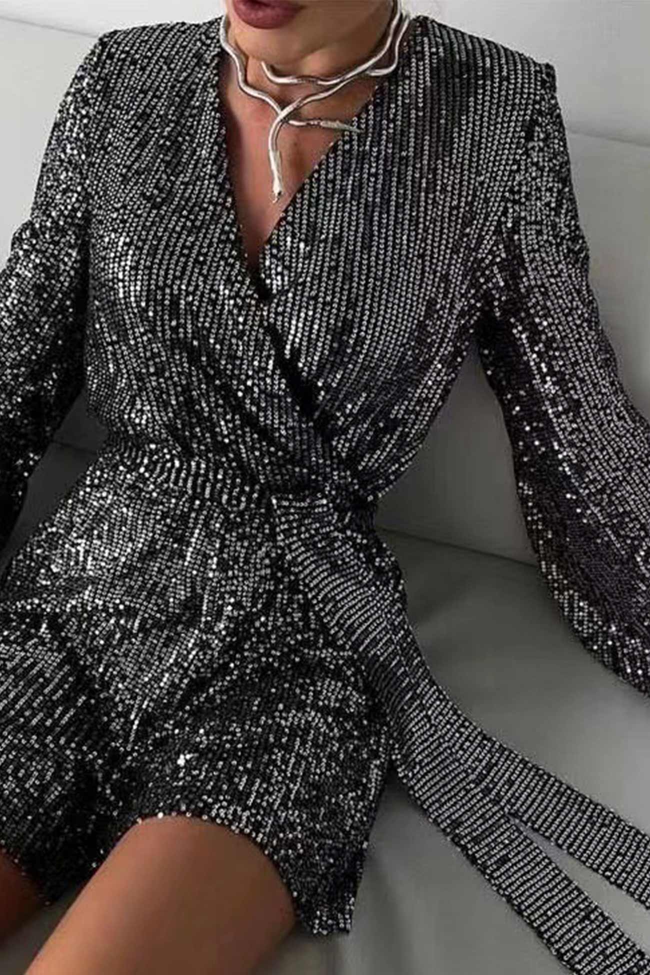 Kvinde iført sort glitrende pailletkjole med dyb V-udskæring og bælte, sidder på grå sofa. Elegant festkjole, moderne mode, glamourøst look.