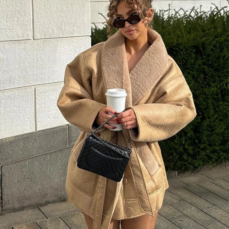 Kvinde iført stor beige frakke, holder en kaffekop og sort quiltet taske. Hun har solbriller på og står foran en hæk. Mode, vintertøj, street style.