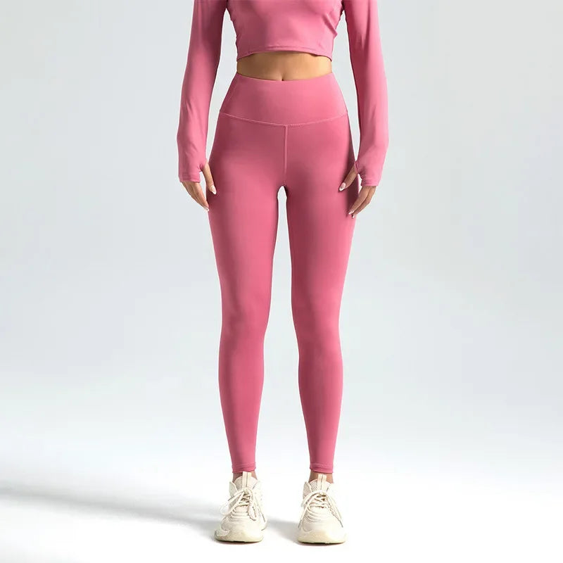 Kvinde iført lyserødt træningssæt med lange ærmer og højtaljede leggings, stående mod en neutral baggrund. Fitnessmode, aktivt tøj, komfortabelt design.