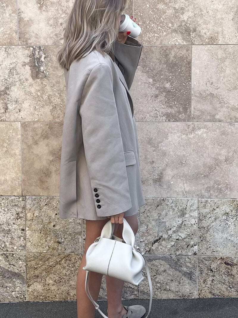 Kvinde i grå blazer drikker kaffe, bærer hvid taske og grå loafers. Står foran beige stenvæg. Mode, street style, minimalistisk outfit.