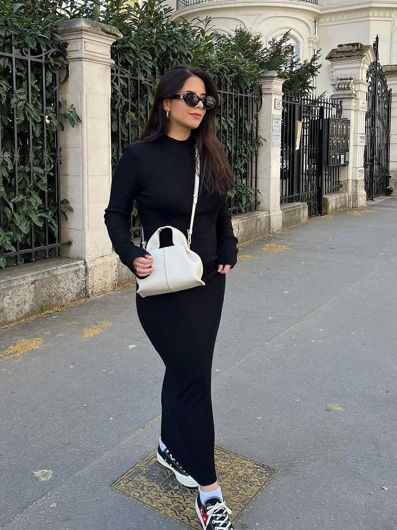 Kvinde i sort kjole og sneakers, bærer hvid taske, går på fortov foran smedejernshegn og historisk bygning. Mode, gade, stilfuld, byliv.