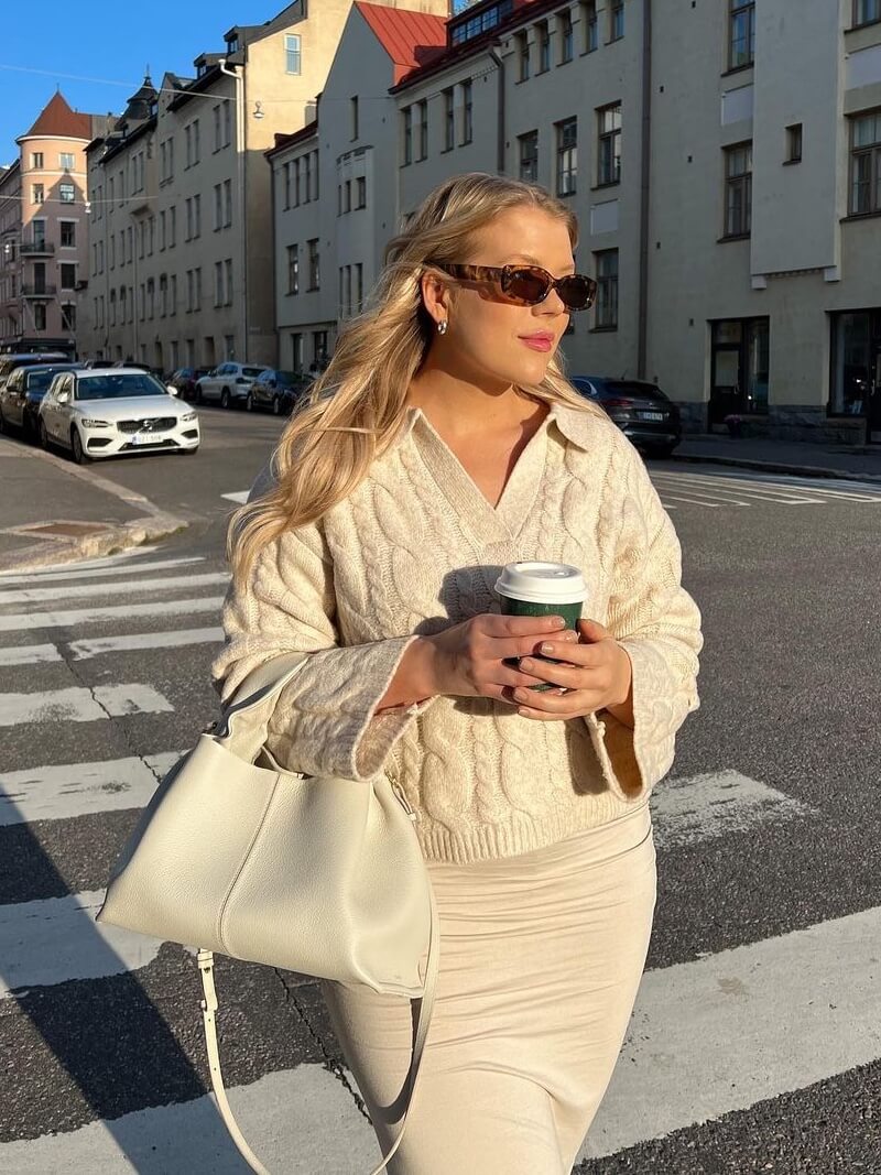 Jeg kan ikke identificere personer på billedet, men her er en passende alt-tekst: Kvinde i beige tøj med solbriller og taske krydser gade i solskin, holder kaffekop.