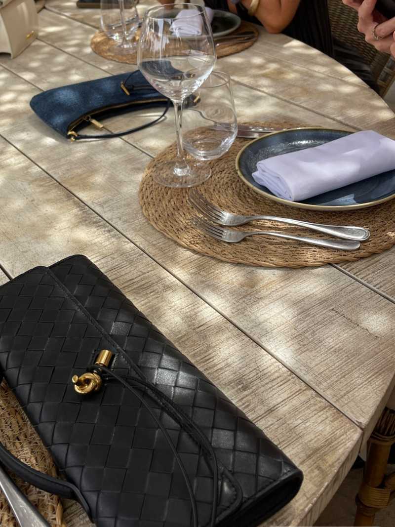 Sort flettet lædertaske på rustikt træbord med dækketallerken, bestik og vinglas. Elegant borddækning udendørs.