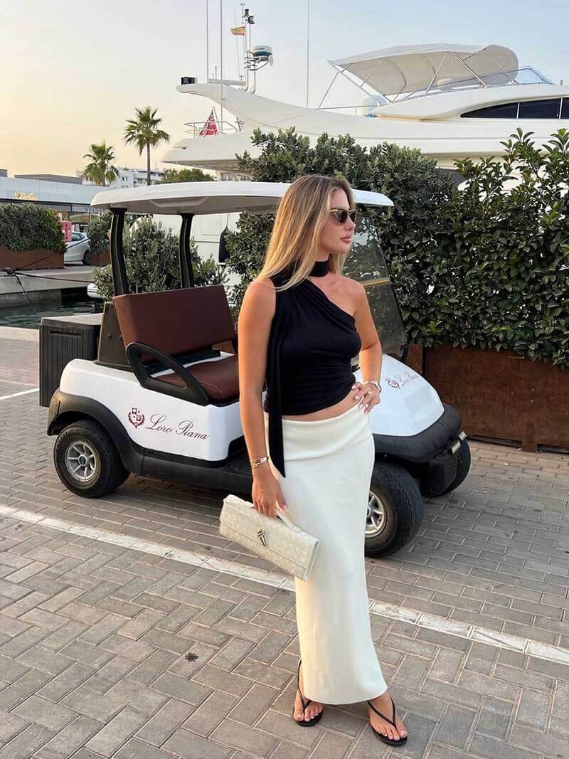 Kvinde i sort top og hvid nederdel står foran golfvogn ved marina. Hun holder en hvid clutch. Luksusyacht og palmer i baggrunden.