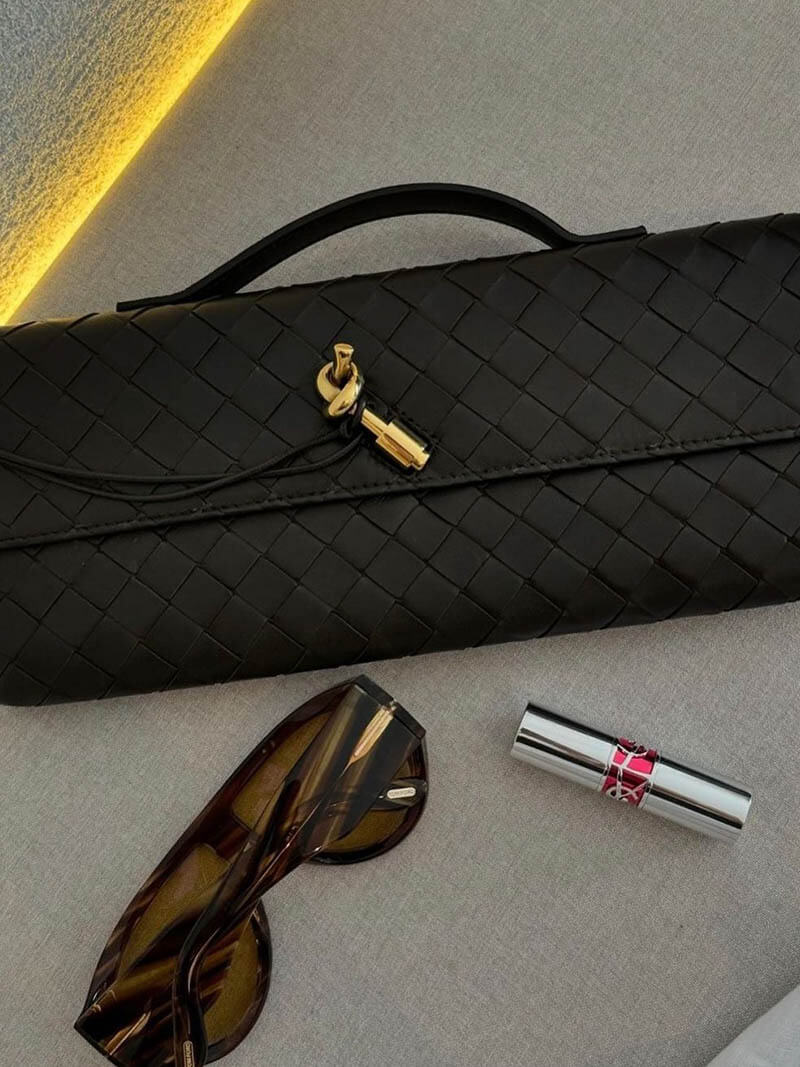 Sort flettet clutch taske med guldspænde, solbriller og læbestift på grå overflade. Stilfuldt tilbehør, mode, luksus, kvindelig elegance.