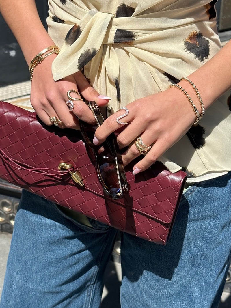 Kvinde med bordeaux fletmønstret clutch, iført guldarmbånd og ringe, holder solbriller. Beige bluse og blå jeans. Modeaccessories udendørs.