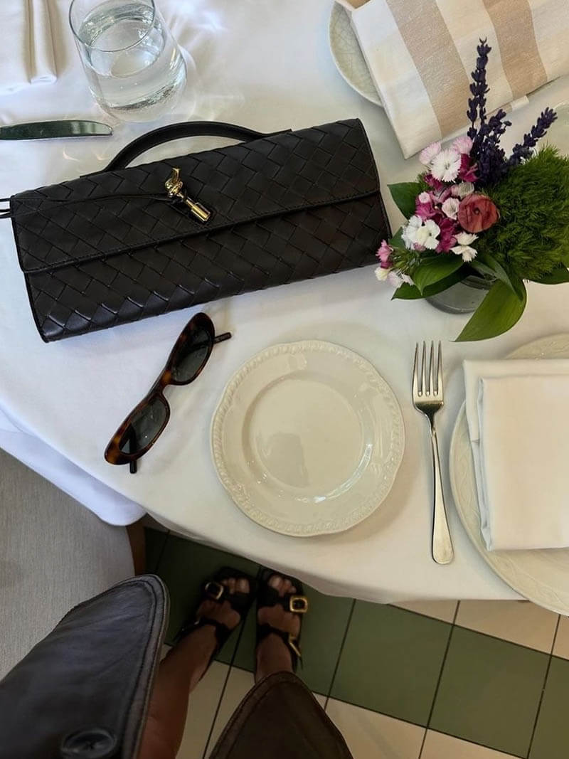 Sort flettet clutch på hvid dug ved siden af tallerken, bestik, solbriller og blomsterbuket. Elegant borddækning, modeaccessories.