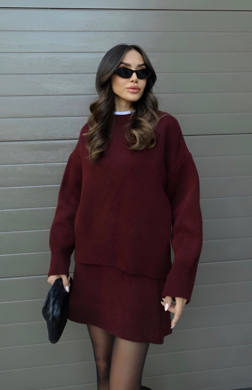 En kvinde iført en bordeaux sweater og nederdel, sorte solbriller og holder en sort taske. Moderne mode, stilfuldt outfit, efterårstøj.