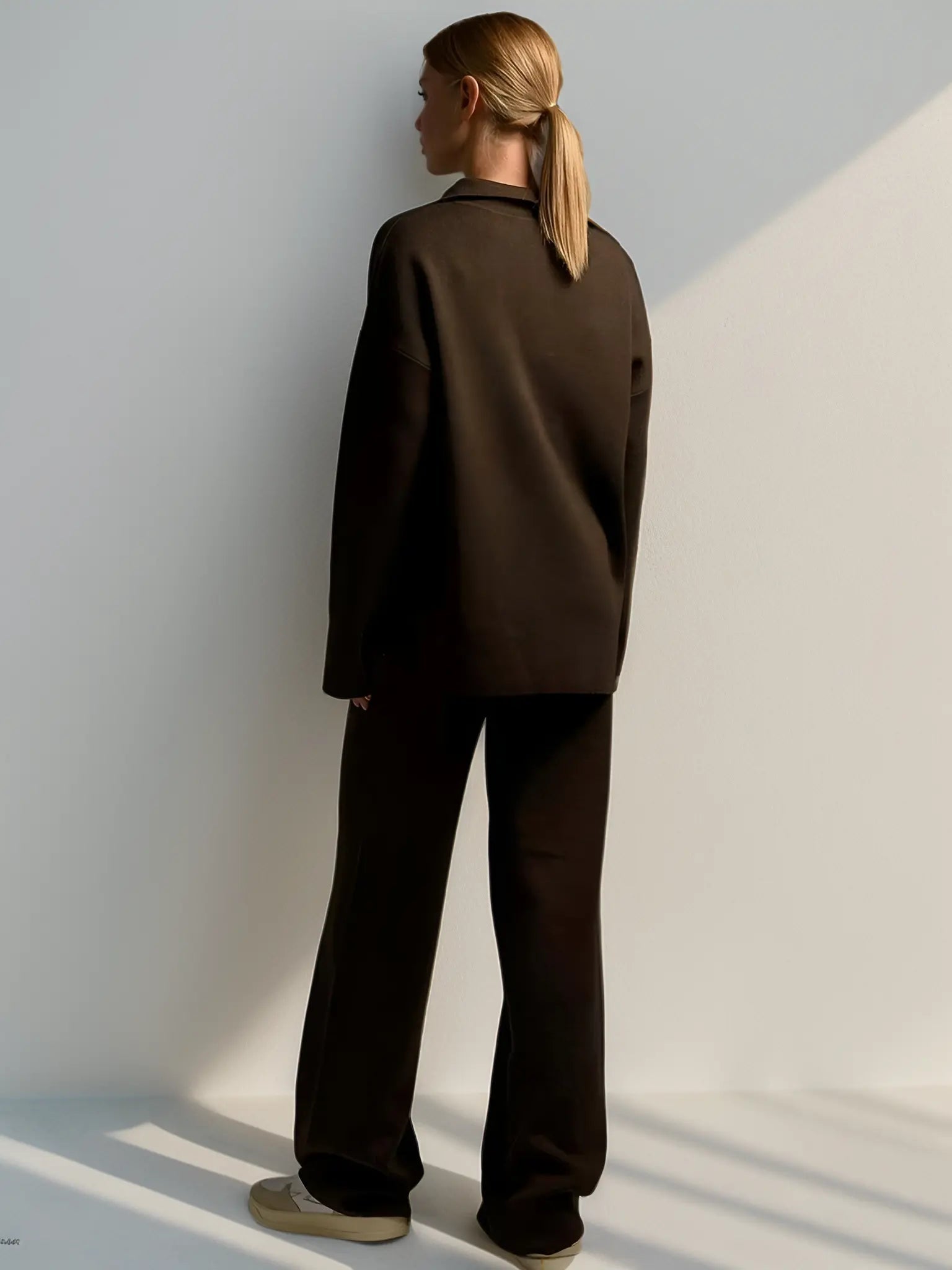 Kvinde i mørkebrun jakkesæt, set bagfra, står mod hvid væg. Minimalistisk mode, elegant stil, moderne kvindetøj, skandinavisk design.