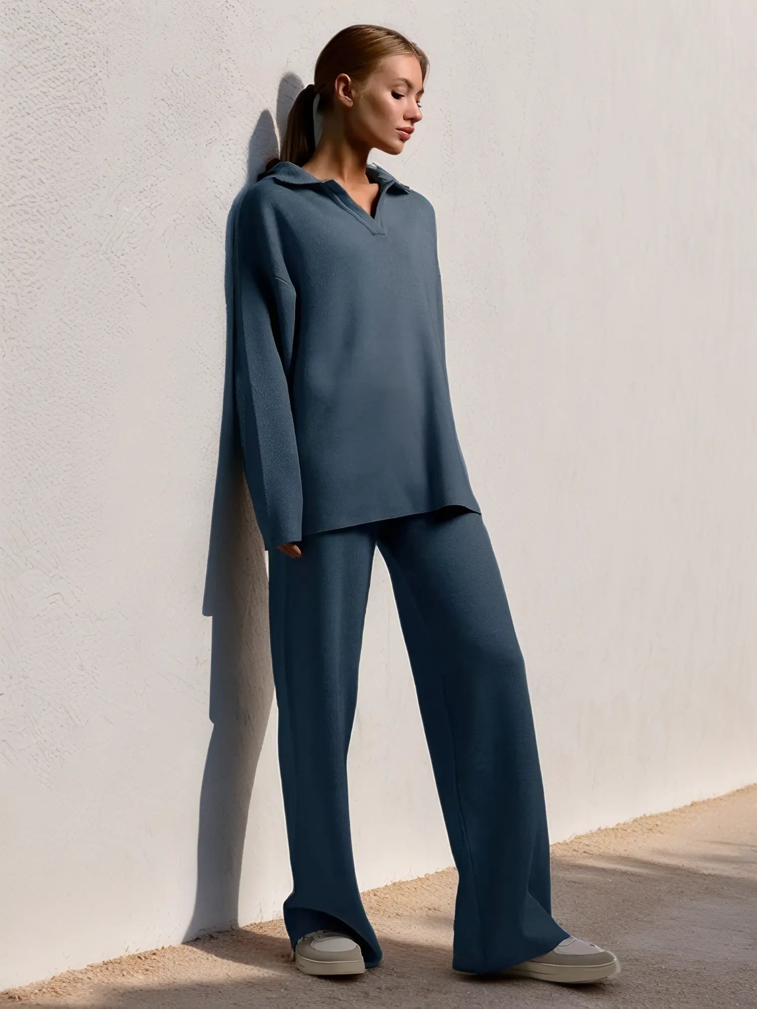 Kvinde iført afslappet, blå lounge-sæt med lange ærmer og bukser, læner sig mod hvid væg. Komfortabelt tøj, moderne stil, afslappet look.