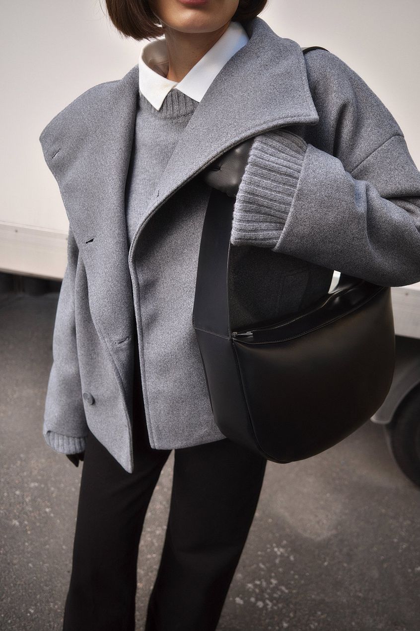 Kvinde iført grå uldfrakke, hvid skjorte og grå sweater, bærer sort lædertaske. Moderne efterårsoutfit, stilfuld og elegant mode.