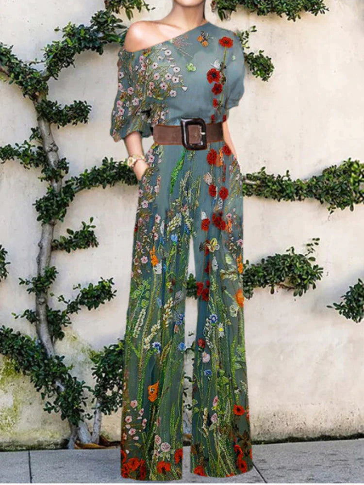 Kvinde iført blomstret buksedragt med bælte, stående foran væg med klatreplanter. Mode, sommerstil, elegant tøj, blomsterprint jumpsuit.