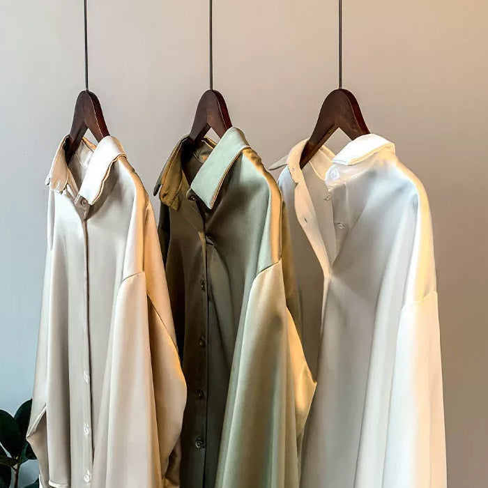 Tre elegante skjorter i beige, olivengrøn og hvid hænger på træbøjler. Perfekt til modebevidste, stilfulde og moderne garderober.