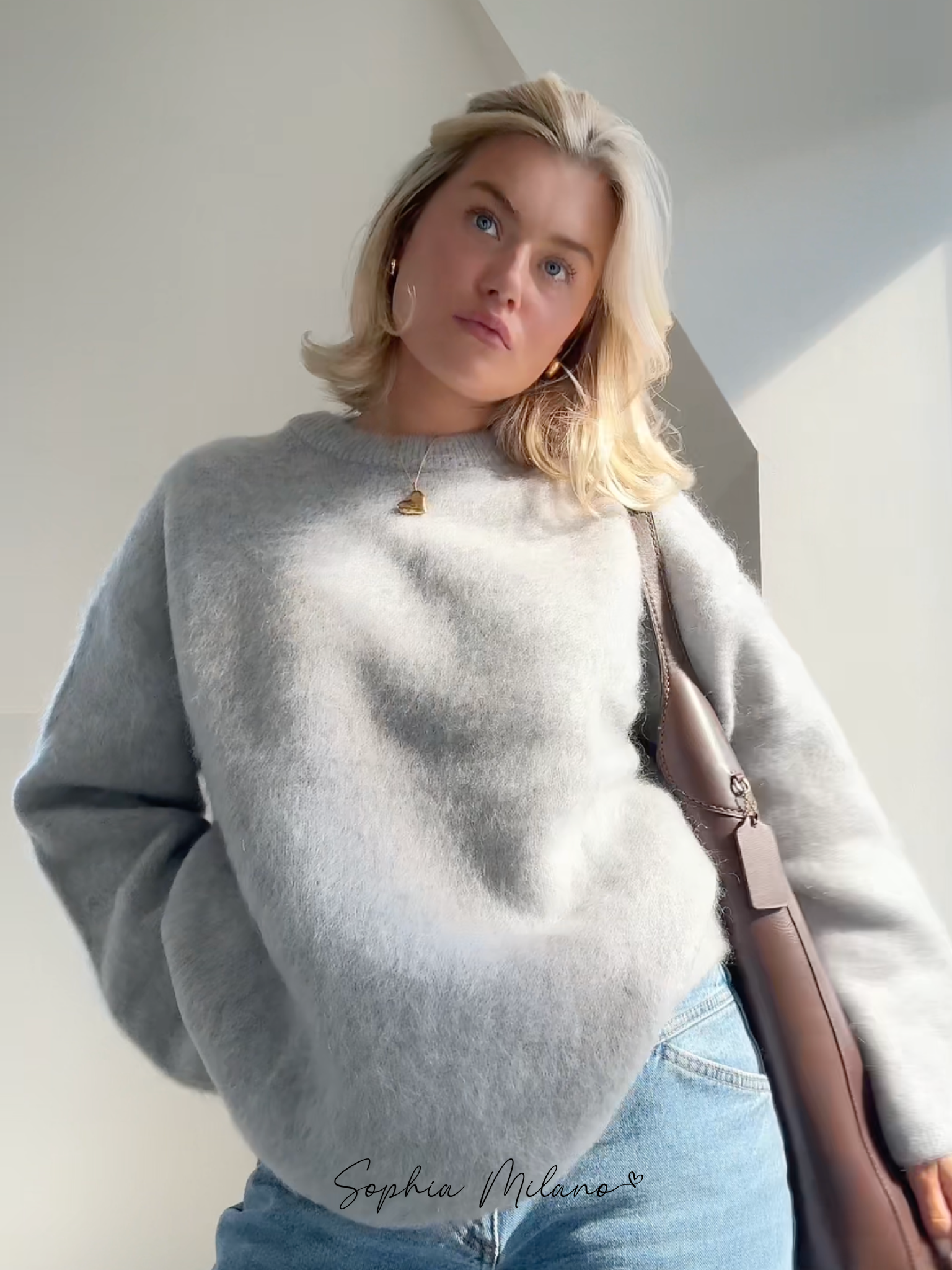 Ung kvinde i grå sweater og jeans, bærer brun taske. Moderne mode, afslappet stil, blondt hår, naturligt lys.