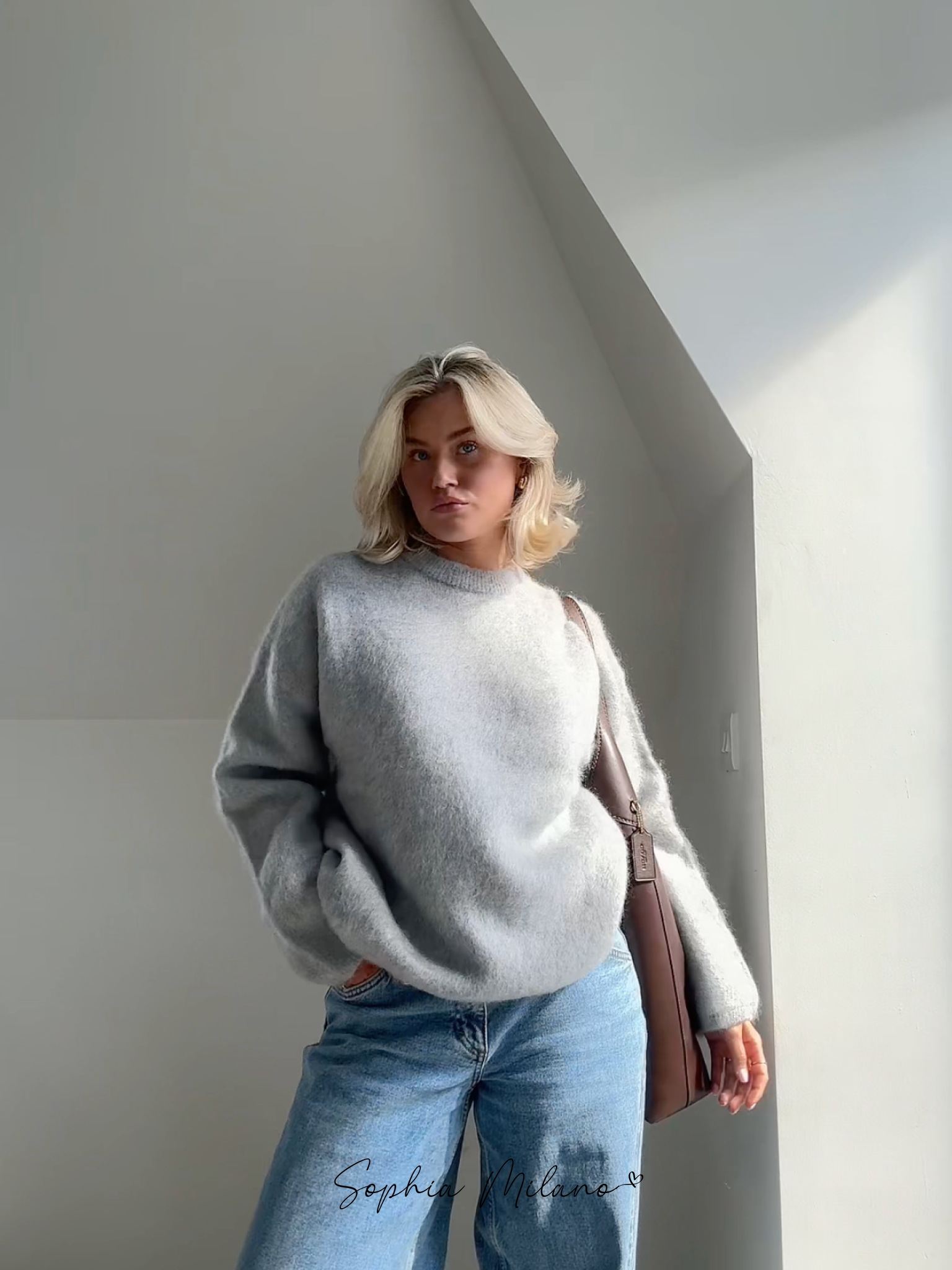 Blond kvinde i grå sweater og blå jeans står i sollys ved hvid væg. Moderne mode, afslappet stil, minimalistisk indretning.